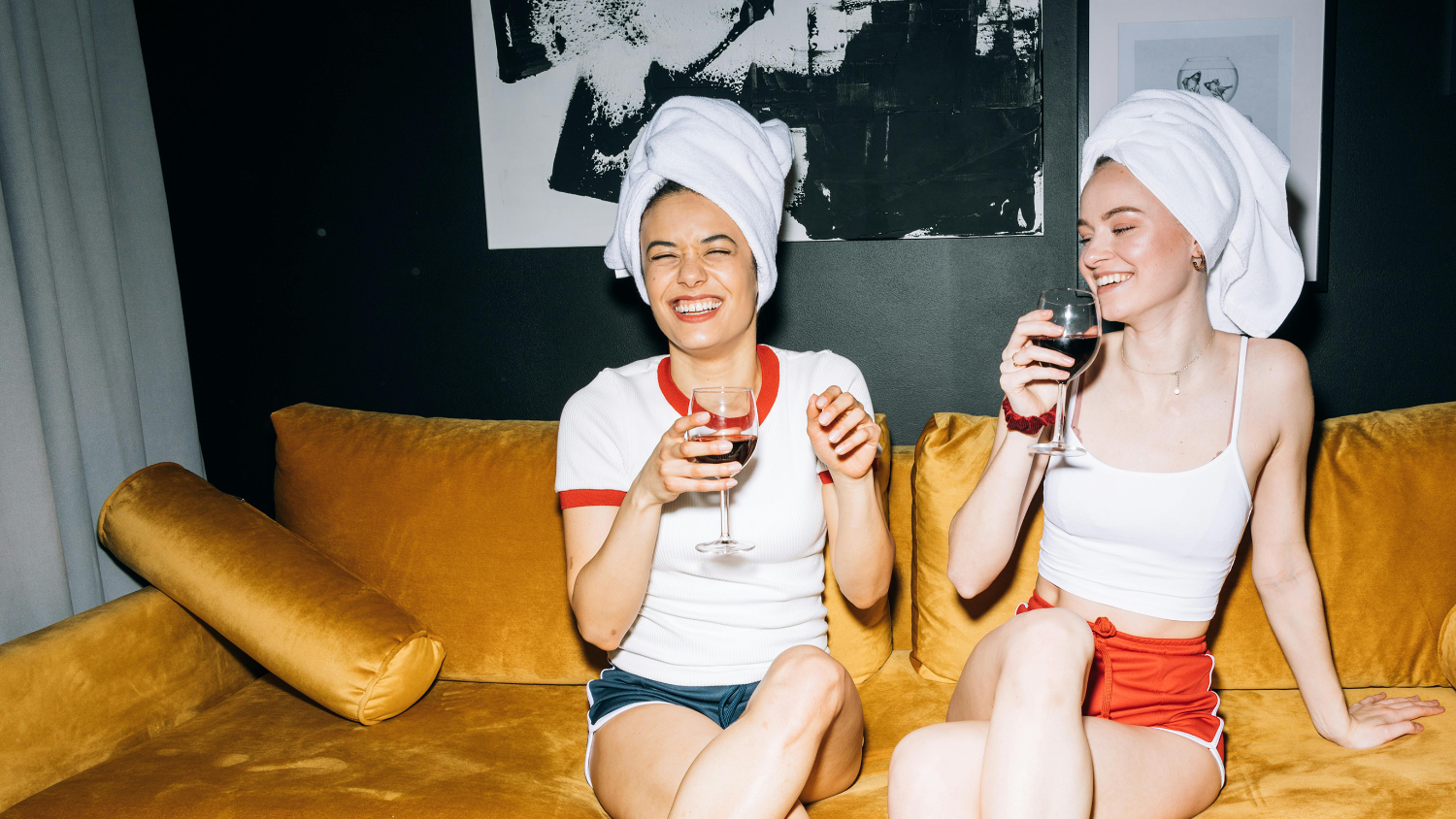 two ladies with towels in their hair happily sipping wine on the sofa